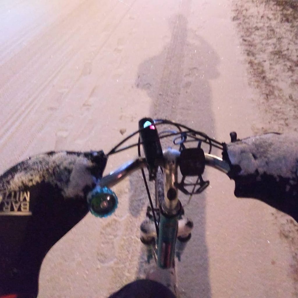 A little blurry but still visible- point of angle is down to show front  of bike with bar mitts covered in snow and frost, left handle bar side  has teal bike bell, front white light on and in the center of handlebar,  right handle bar side has silicone phone holder with no phone in it;  background/below the bike is the Greenway covered in snow with tire  tracks and shoe prints to the left of the bike.