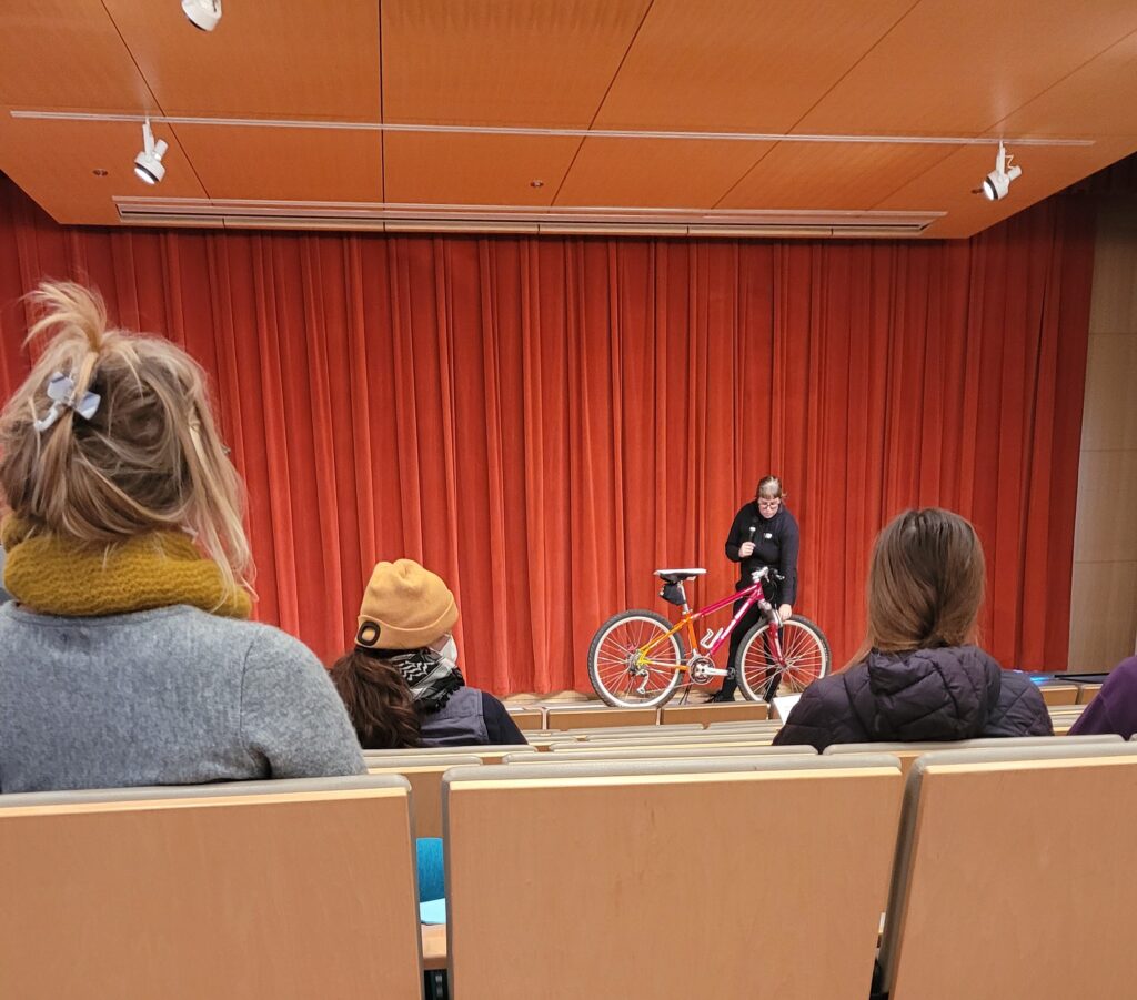 A person on a stage with a bike pointing to different parts of the bike.