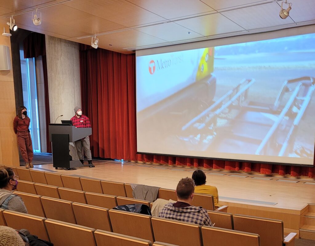 Two people stand on stage looking at a projected image of a bike rack on the front of a bus.