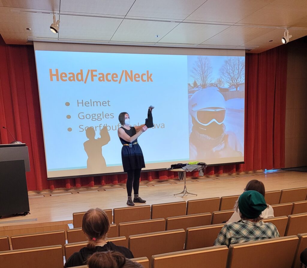 Woman holds up a face buff on a stage in front of a projected slide that says "Head/Face/Neck: Helmet, googles, scarf, buff, balaclava". The slide shows a person wearing a helmet, goggles, and a warm balaclava on her face.