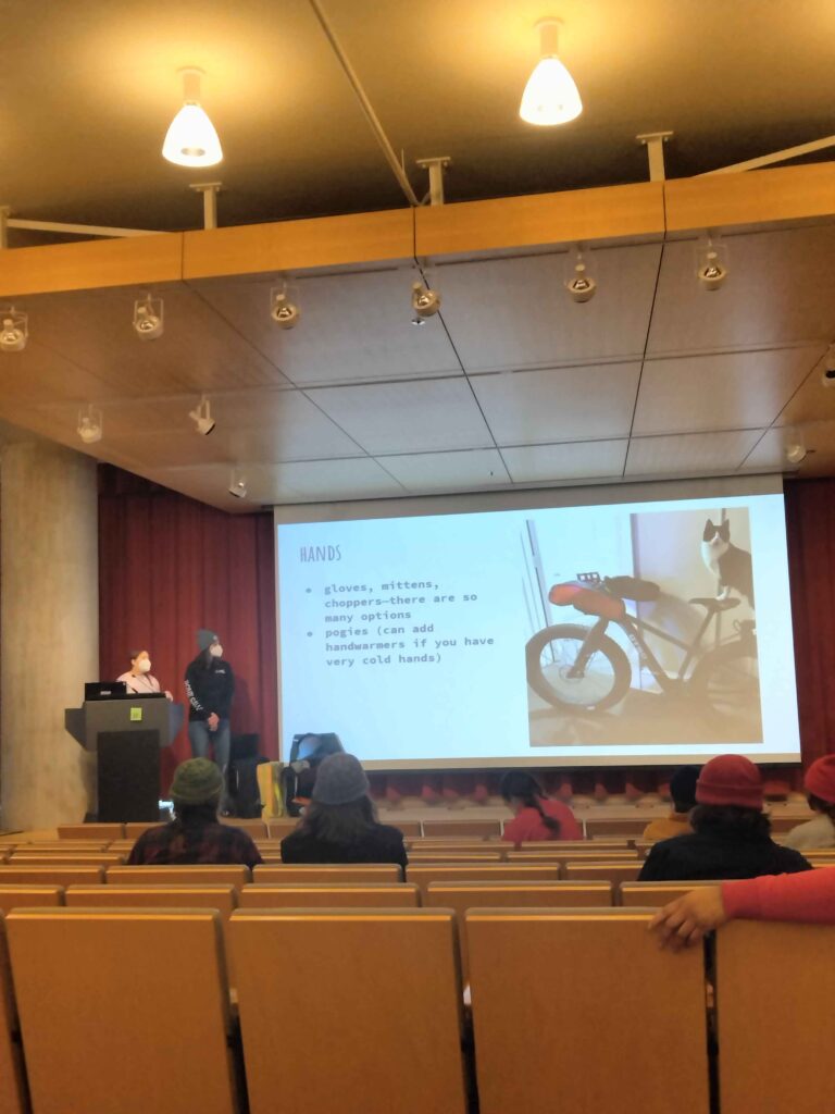 Stage with two people presenting. Slide shows ideas for things to wear to keep your hands warm, and a photo of a cute cat standing on a bicycle seat.
