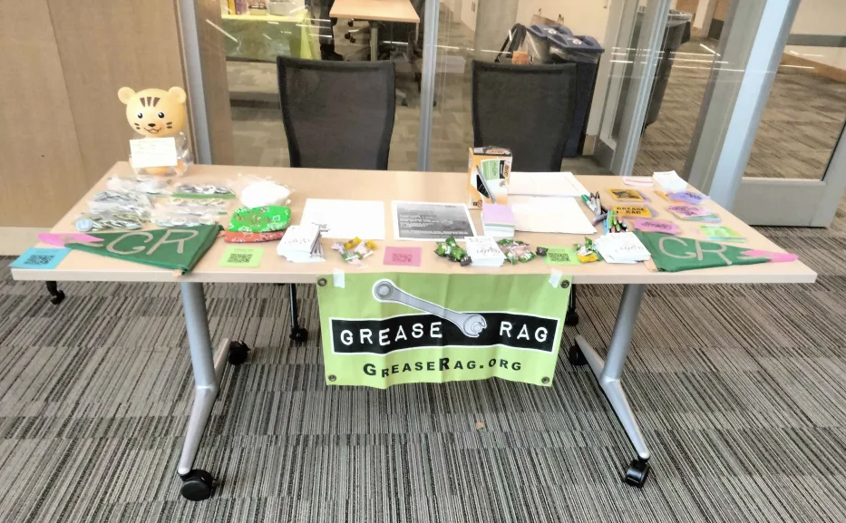 Grease Rag welcome table with flyers, candy, teddy bear donation jar, pens, colorful banners, and spoke cards.