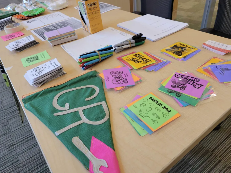 Closeup of Grease Rag welcome table with hand-sewn banner, pens, stacks of zines, and colorful spoke cards.